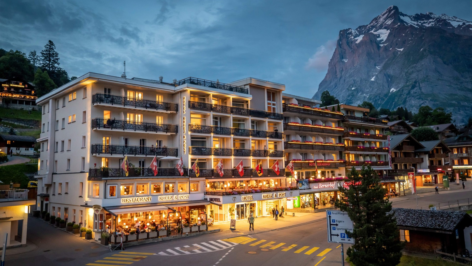 Dusk view of a bustling hotel with warmly lit balconies and Swiss flags, set against towering mountains, exuding a cozy yet vibrant retreat for travelers.
