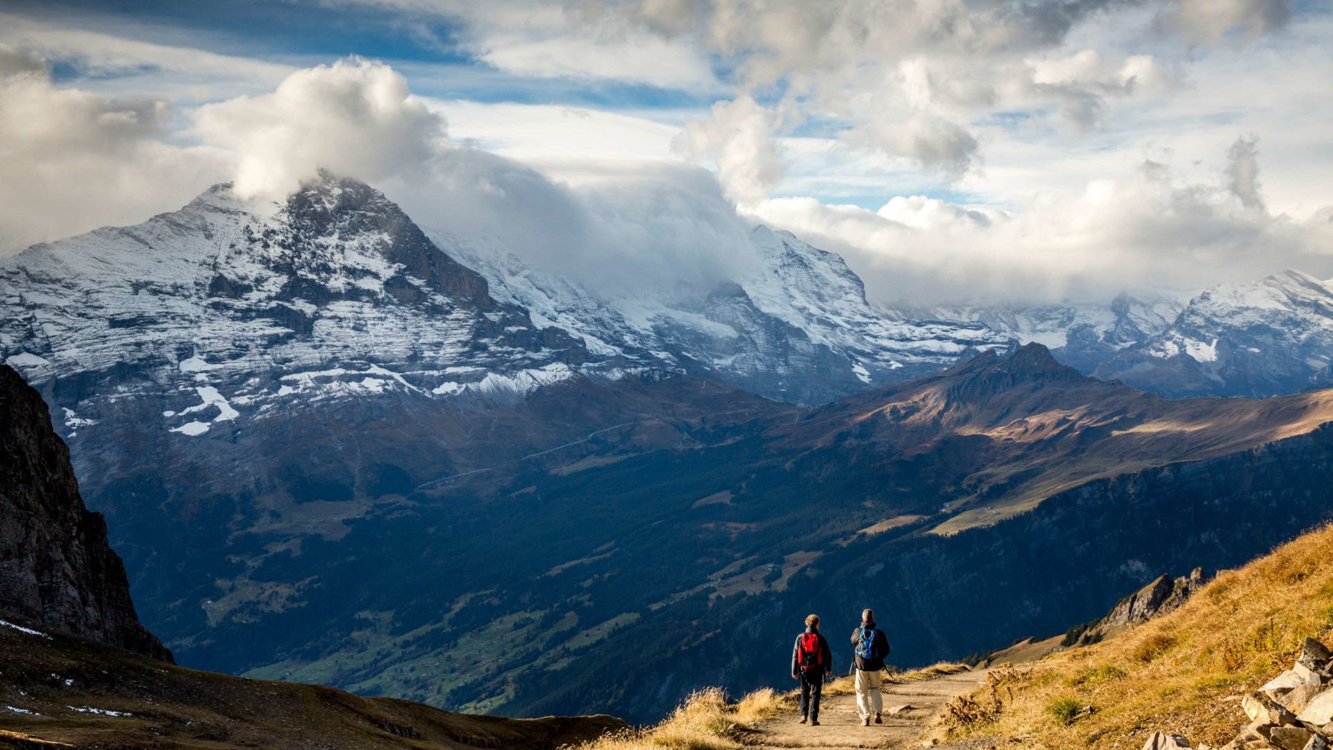 Two hikers on a scenic trail with snow-capped mountains and vast clouds, evoking a sense of adventure and tranquility in nature.