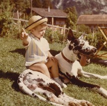 Vintage photo of a young boy in a straw hat and striped shirt sitting next to a large harlequin Great Dane in a sunny, grassy backyard, capturing a moment of cheerful companionship.