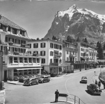 Vintage black-and-white photograph of a bustling hotel plaza with classic cars, framed by dramatic mountain peaks, evoking a nostalgic and serene ambiance for travel enthusiasts.