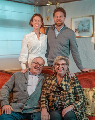 Four people of varying ages smiling warmly in a cozy, festively decorated living room, reflecting a multi-generational family connection.