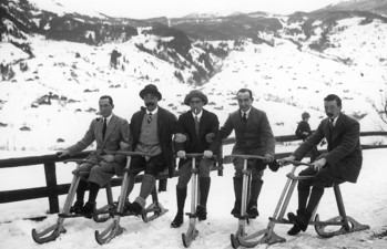 Five men in vintage clothing on wooden sleds against a snowy mountain backdrop, evoking a nostalgic and adventurous spirit in winter sports history.