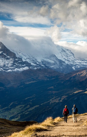 Two hikers explore a majestic mountain trail, amid towering snow-capped peaks and expansive blue skies, capturing the essence of adventure travel.