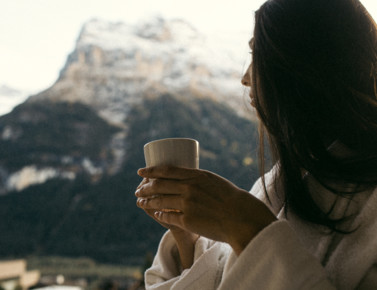 Woman in a cozy robe enjoying a warm drink, serene mountain backdrop, evoking tranquility and relaxation in a natural setting.