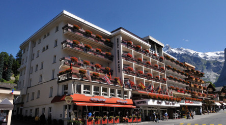 Alpine hotel with vibrant red flower boxes on balconies, set against a backdrop of towering mountains, projecting a serene and inviting retreat for travelers.