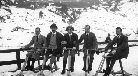 Five men in vintage clothing on wooden sleds against a snowy mountain backdrop, evoking a nostalgic and adventurous spirit in winter sports history.