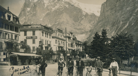Vintage sepia-toned photo of cyclists in early 20th-century attire in front of majestic mountain hotels, evoking a nostalgic and serene atmosphere ideal for history buffs.
