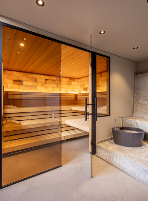 Warm wooden sauna with reflective glass door beside minimalist grey stone basins, exuding tranquility for wellness spaces.
