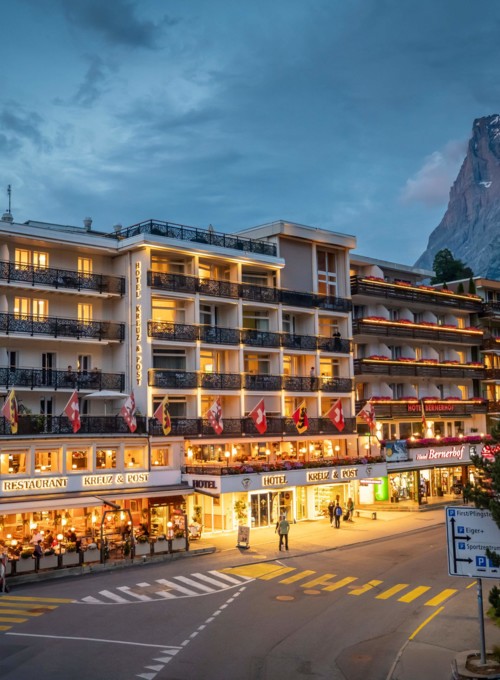 Dusk view of a bustling hotel with warmly lit balconies and Swiss flags, set against towering mountains, exuding a cozy yet vibrant retreat for travelers.