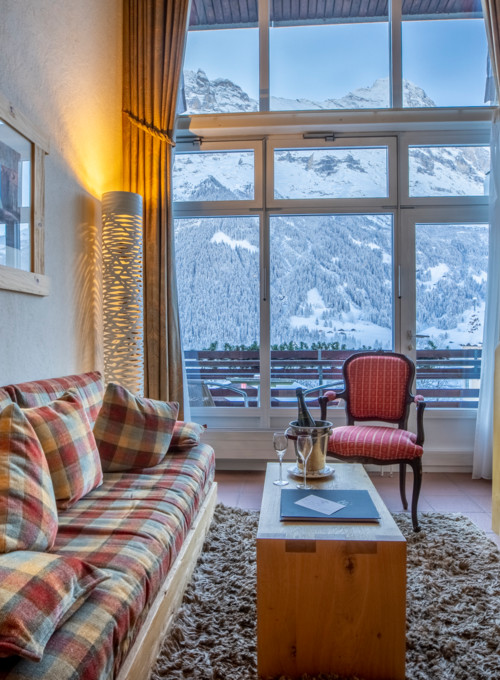 Cozy alpine living room with plaid sofa and large window overlooking snowy mountains, offering a warm and serene escape for vacationers.