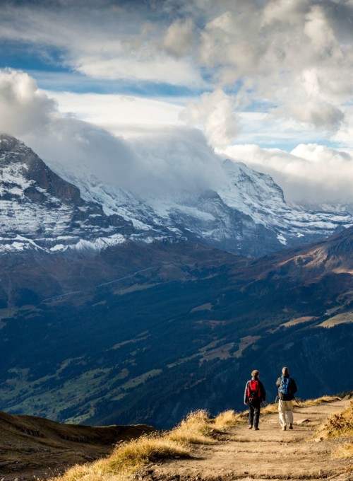 Two hikers on a scenic trail with snow-capped mountains and vast clouds, evoking a sense of adventure and tranquility in nature.