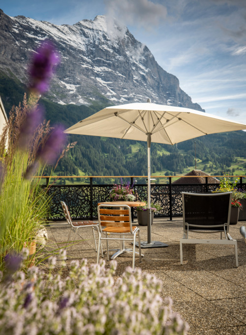 Alpine terrace with a beige umbrella and wooden furniture, set against a majestic mountain backdrop, evoking serenity perfect for travel and leisure.