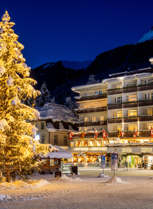 Snow-covered Christmas tree illuminated by golden lights against a backdrop of quaint, festive hotels, evoking a cozy, welcoming winter ambiance ideal for holiday tourism.