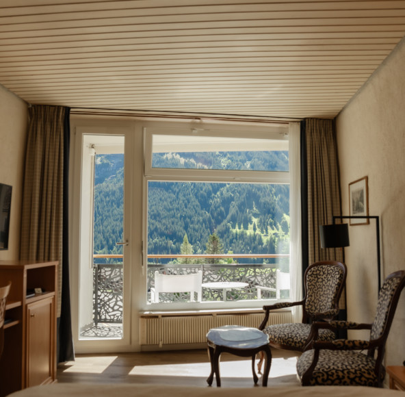 Cozy hotel room with a wooden floor, elegant furniture, and a balcony overlooking lush mountains, creating a serene and inviting retreat for travelers.