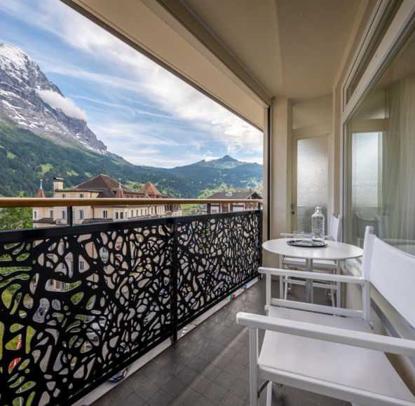 Alpine balcony view with intricate metal railing, white furniture against majestic mountains, offering a serene retreat for travel and relaxation.