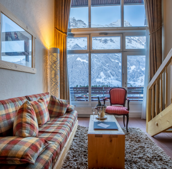 Cozy alpine living room with plaid sofa and large window overlooking snowy mountains, offering a warm and serene escape for vacationers.