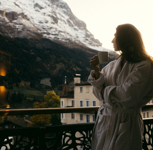 Woman in white robe enjoys coffee on a balcony, serene mountain backdrop, inspiring tranquility and leisure in travel.