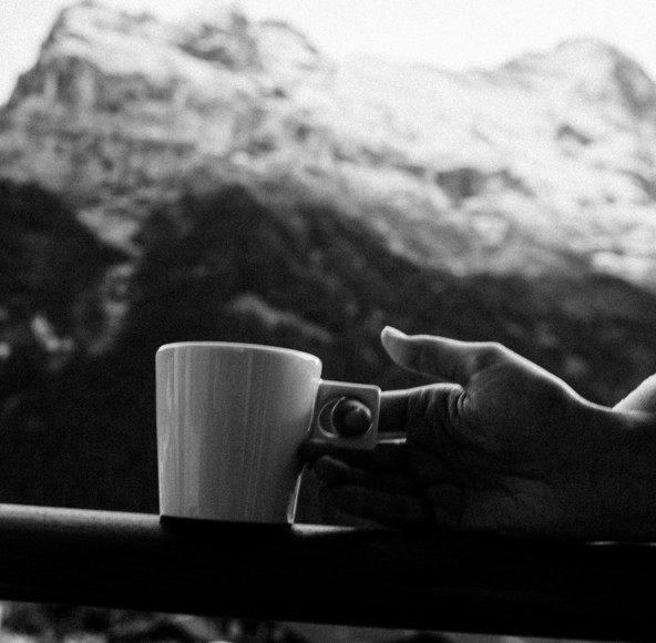 Monochrome photo of a hand holding a white coffee cup with serene mountain backdrop, expressing tranquility for relaxation seekers.