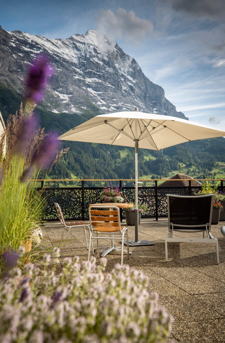 Alpine terrace with umbrella and seating, set against towering mountains, offering serenity and breathtaking views for travel and leisure.