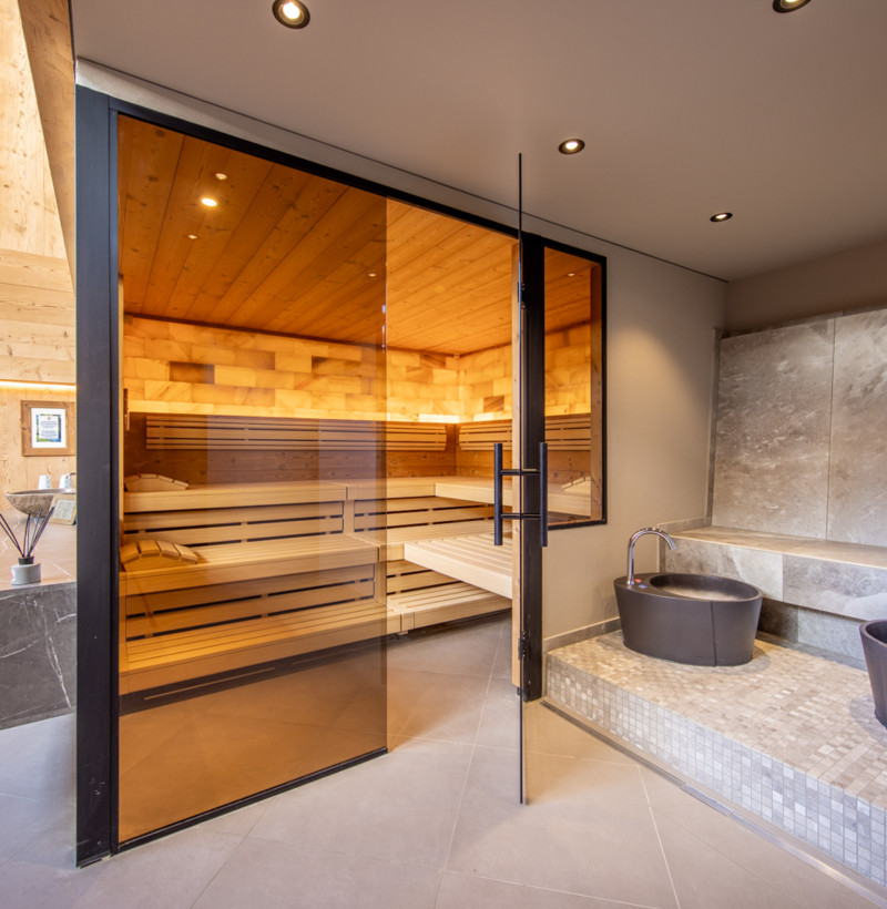 Warm wooden sauna with reflective glass door beside minimalist grey stone basins, exuding tranquility for wellness spaces.