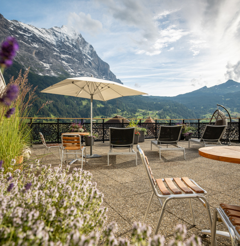 Alpine terrace with chairs and umbrella, framed by majestic mountains, creating a serene getaway spot for travel and leisure.