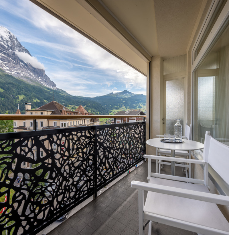 Alpine balcony view with intricate metal railing, white furniture against majestic mountains, offering a serene retreat for travel and relaxation.