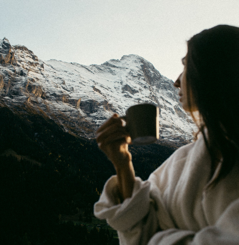 Misty morning view of snow-capped mountains enjoyed by a woman holding a warm mug, evoking serenity and peace, perfect for leisure and travel-themed promotions.