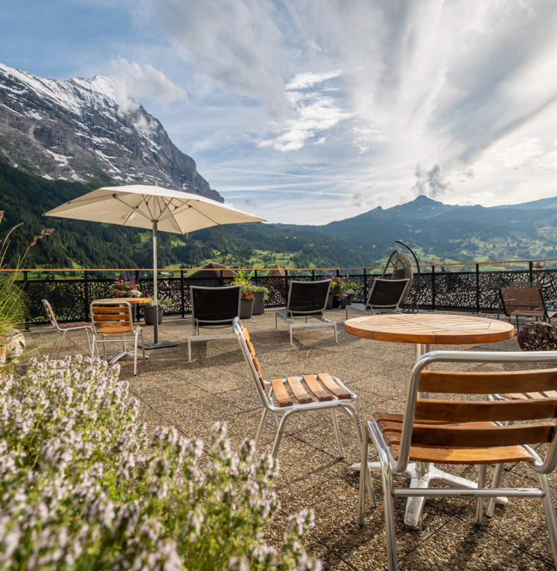 Alpine terrace with cozy seating and lush greenery, set against a backdrop of majestic mountains, evoking tranquility and escape for travel enthusiasts.