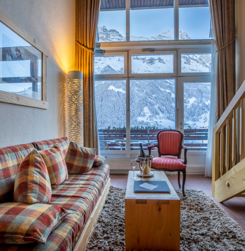 Cozy alpine living room with plaid sofa and large window overlooking snowy mountains, offering a warm and serene escape for vacationers.