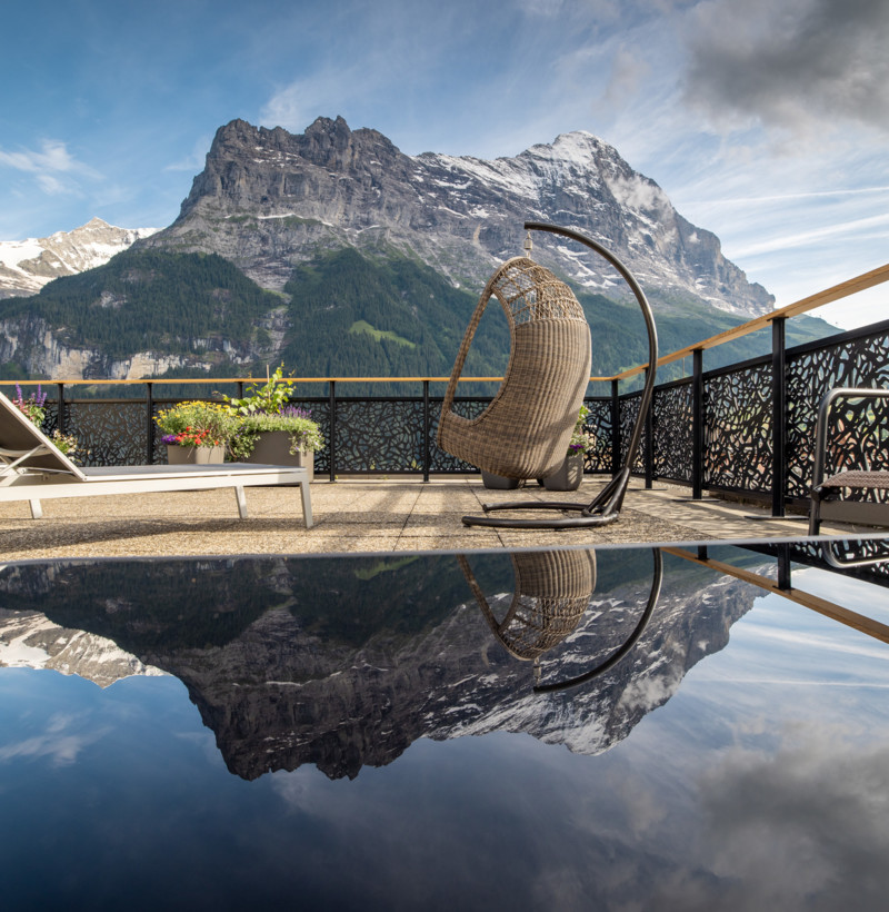 Tranquil terrace with a woven egg chair and reflective water surface, framed by majestic mountains, evoking serenity in a luxurious setting.