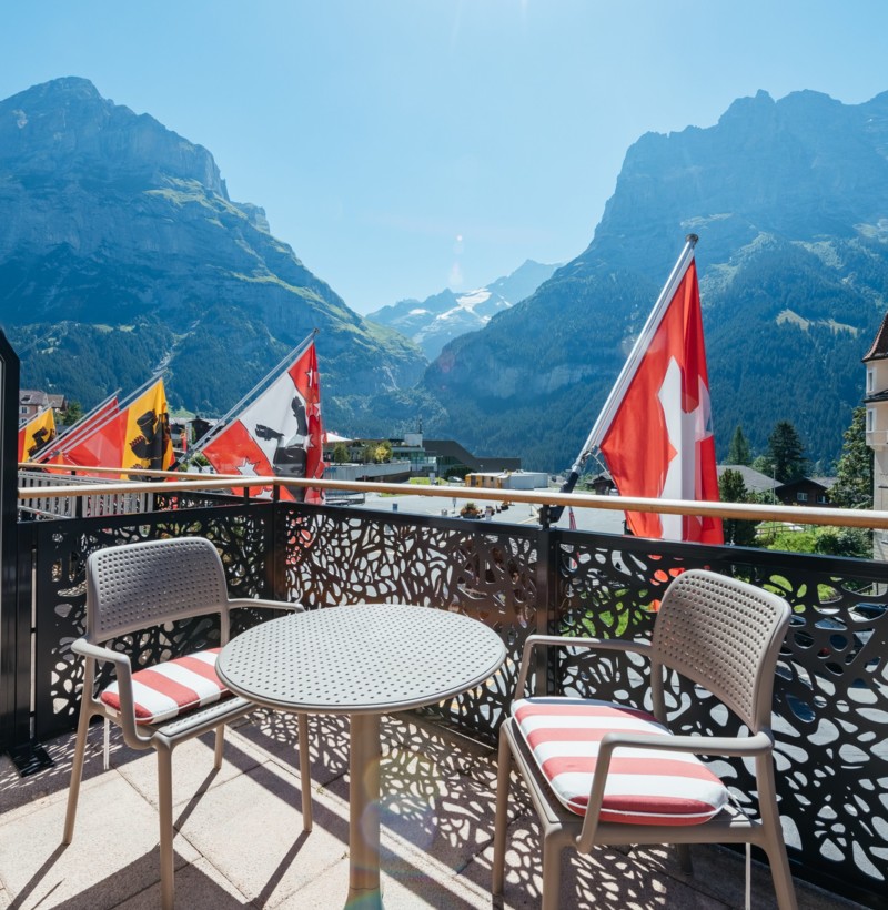 Sunny balcony with Swiss flags, mountain views, and comfortable seating, ideal for relaxation and tourism.