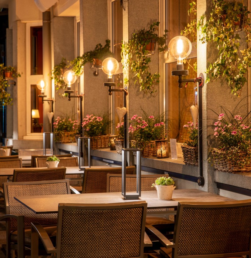 Warmly lit outdoor café at night with wicker chairs and blooming plants, exuding a cozy and inviting ambiance for dining.