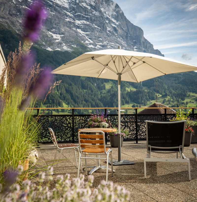 Alpine patio setting with a sunlit umbrella and wooden furniture, evoking tranquility against a mountain backdrop, ideal for travel and leisure sectors.