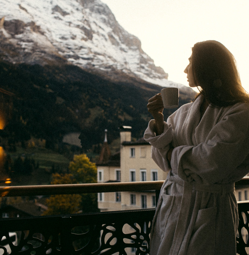 Woman in white robe enjoys coffee on a balcony, serene mountain backdrop, inspiring tranquility and leisure in travel.