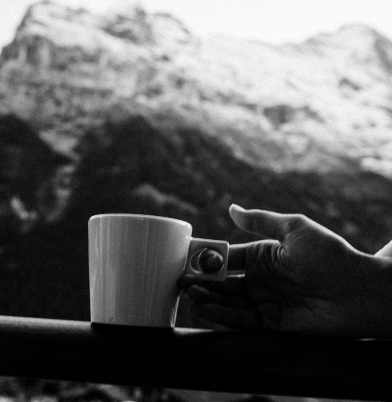 Monochrome photo of a hand holding a white coffee cup with serene mountain backdrop, expressing tranquility for relaxation seekers.