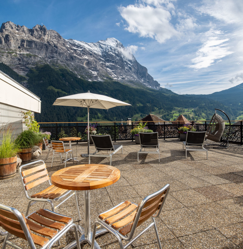 Alpine terrace with wooden furniture and umbrella, set against majestic mountains, offers a serene, inviting ambiance for relaxation and tourism.