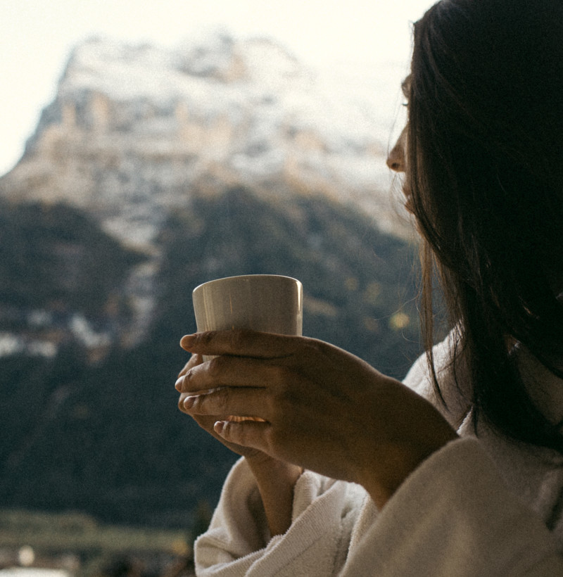 Woman in a cozy robe enjoying a warm drink, serene mountain backdrop, evoking tranquility and relaxation in a natural setting.