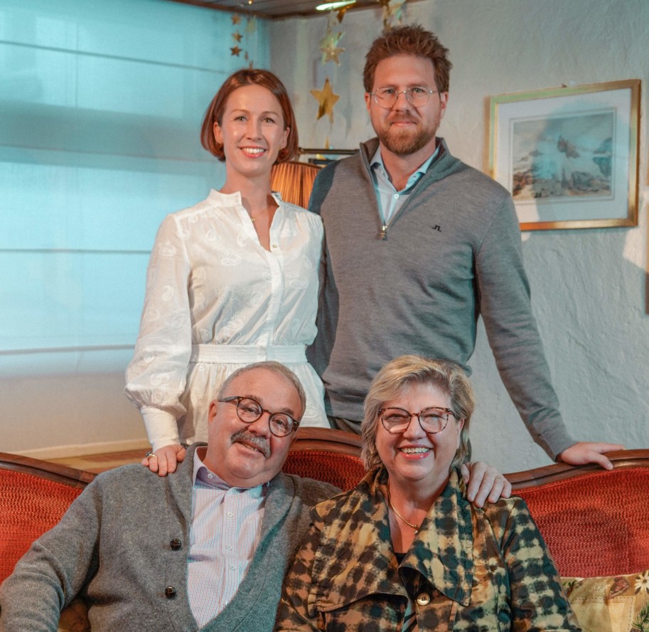Four people of varying ages smiling warmly in a cozy, festively decorated living room, reflecting a multi-generational family connection.